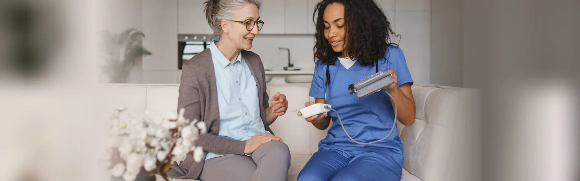 Nurse showing a blood pressure monitor to a smiling senior woman on a couch
